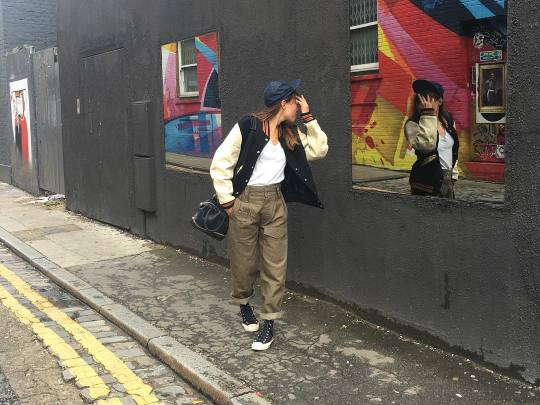 Emilie Casiez, wearing a varsity jacket with fatigue pants, a ball cap and canvas sneakers, checking herself out in a mirrored street window.