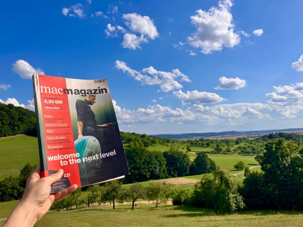Chris holding an old edition of a German Mac magazine in front of rolling green fields and a blue sky.