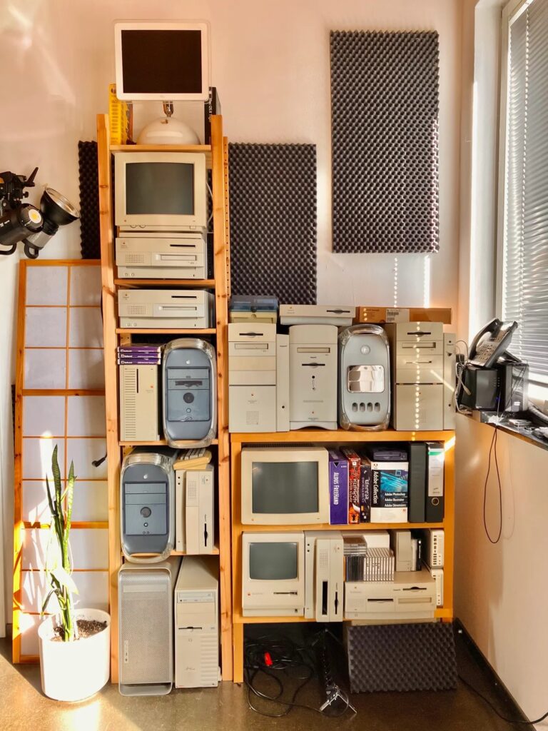 A stack of vintage Mac computers on shelves against a wall, illuminated by sunlight through blinds.
